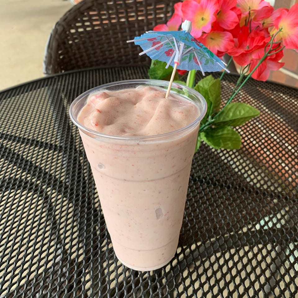 A Capri's Pizzeria strawberry smoothie with a blue umbrella on a black mesh table with red flowers.