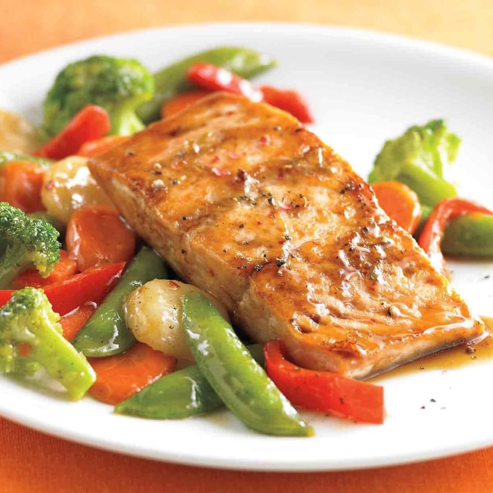 Glazed salmon fillet with broccoli, carrots, red peppers, and snap peas on a white plate.