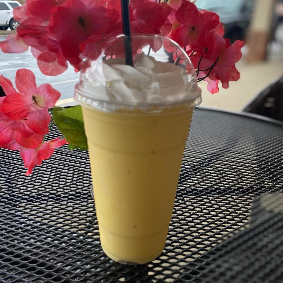 A yellow Capri's Pizzeria smoothie with whipped cream and a straw sits on a black mesh table.